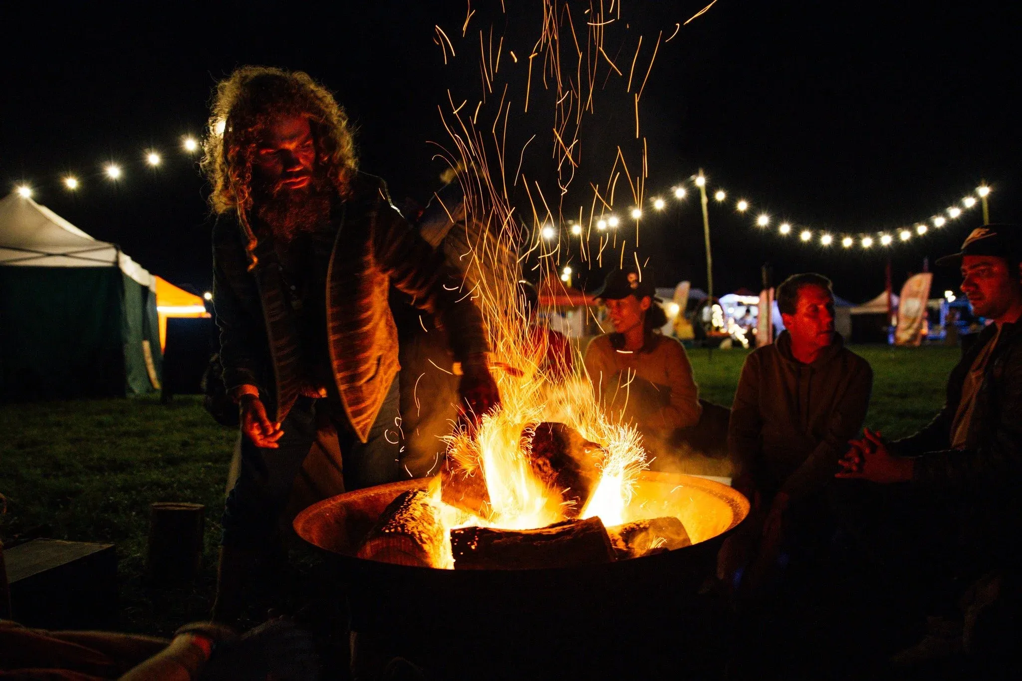 Guests gathered around a fire bowl under warm festoon lighting at a night festival campsite